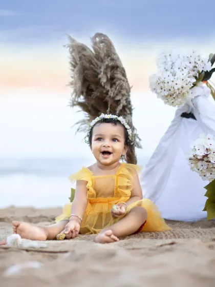 A baby girl in a yellow dress laughs with delight during her first birthday cake smash session on the beach, surrounded by sand and waves.