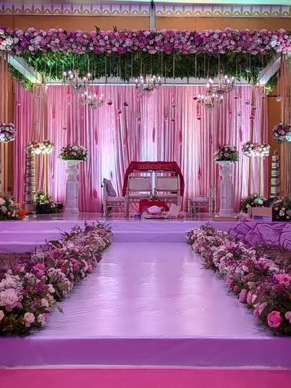 A fairytale aisle for a wedding ceremony. The path is lined with lush arrangements of pink and purple flowers, leading to a romantic stage draped in soft pink fabrics.