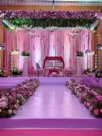 A beautiful mandap setup with a floral-lined aisle leading to a stage draped in soft pink fabric. The canopy of flowers and greenery adds a touch of romance.