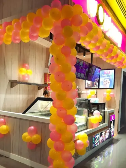 A custom balloon arch designed to fit the structure of a food kiosk for its opening day. The bright pink and yellow colors are cheerful and attract customers.