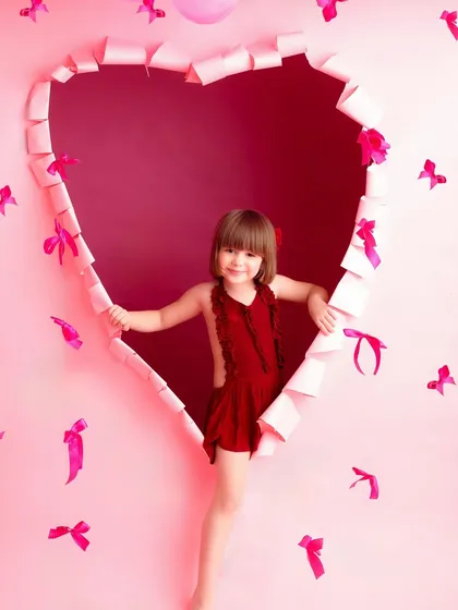 A little girl in a red dress peeks through a heart-shaped cutout in a pink wall. This setup is perfect for a Valentine's Day mini-session, creating a sweet and loving portrait.