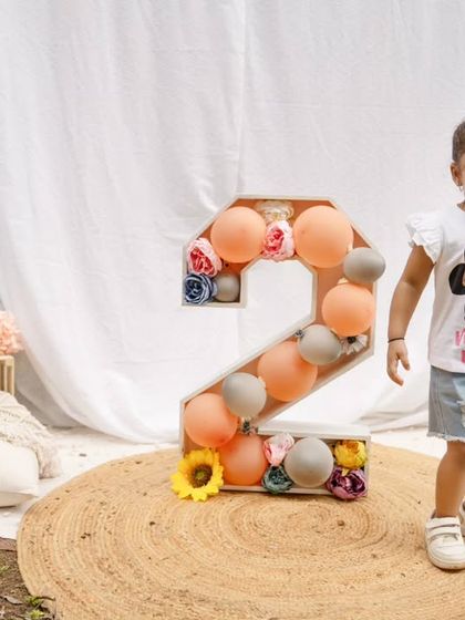 A happy toddler girl stands next to a large number "2" filled with balloons during her outdoor birthday session.