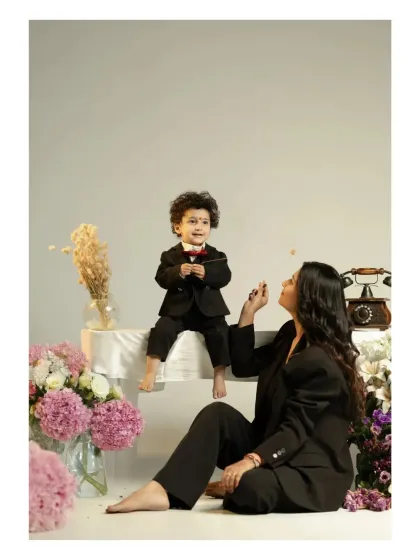 A playful and stylish studio shot. The little boy, dressed in a tiny suit, sits on a table while his mother engages with him from below. The scene is filled with charm, flowers, and a sense of fun.