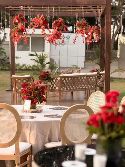 An outdoor sangeet and dinner setup in a garden, with a wooden pergola decorated with hanging red floral arrangements.