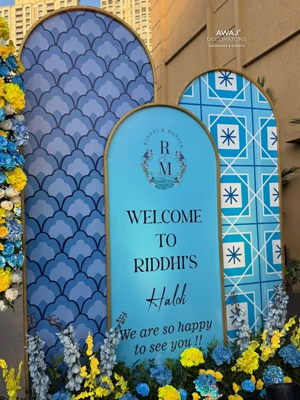 A personalized welcome sign for the bride's Haldi, featuring a custom monogram and surrounded by cheerful yellow and blue florals.