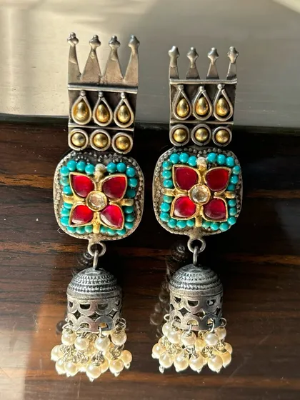 These fusion jhumkas are fit for royalty. They feature a crown-like top with intricate turquoise and red stone work, finished with a classic silver jhumka drop adorned with pearls.