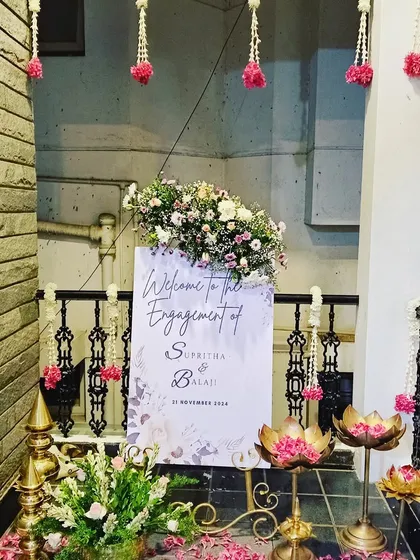 A charming welcome sign for an engagement ceremony. The sign is surrounded by traditional lotus-shaped brass lamps and hanging floral strings, creating a warm and inviting entrance for the guests.