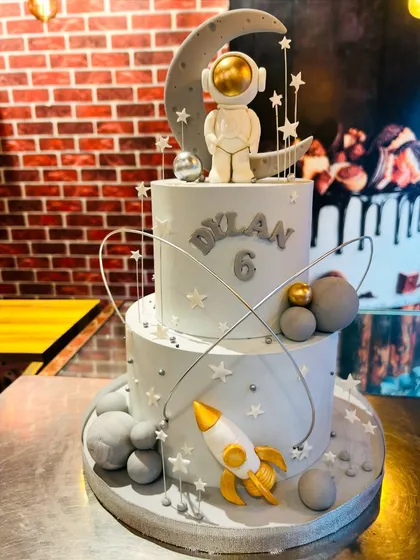 An out-of-this-world two-tier astronaut cake for a 6th birthday. This space-themed cake features a fondant astronaut, a crescent moon, planets, and a rocket ship, creating a galactic adventure for the birthday boy.