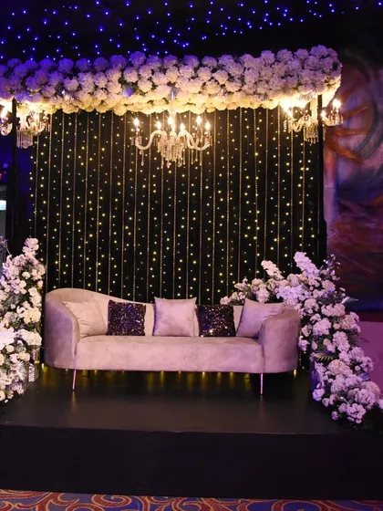 This elegant photo spot features a plush sofa against a curtain of fairy lights, framed by white florals and a classic chandelier, perfect for a romantic Sangeet.