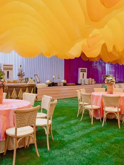 Guest seating at the equestrian Haldi, with wicker chairs and pastel pink tables set on an artificial lawn under the dramatic orange fabric ceiling.