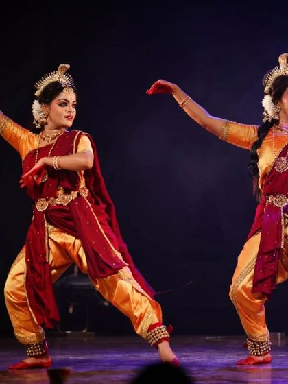 A dynamic pose from "Shrishti Stithi Laya". The dancers' powerful leg extensions and synchronized mudras capture the fierce energy of the cosmic dance. The matching red and gold costumes create a visually unified and striking look.