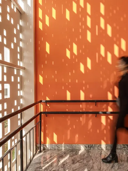 A person walks along a corridor at Vidyashilp Academy, where sunlight filters through the perforated facade, casting patterns on a vibrant orange wall. This image captures the sensory experience of the architecture, where light itself becomes a material.