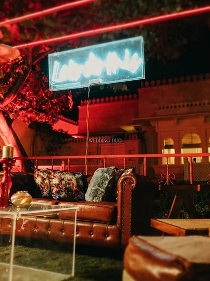A cozy lounge corner for the cocktail night, featuring a classic leather sofa set against a backdrop of red-lit architecture and a modern neon sign, creating a bold and inviting atmosphere.