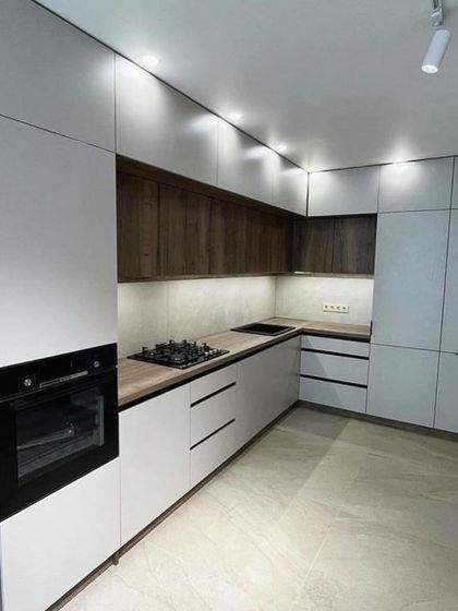 A wide shot of the completed kitchen in Vasant Kunj, featuring a harmonious blend of white and wood-textured cabinets. The L-shaped layout is complemented by bright ceiling spotlights and under-cabinet lighting.