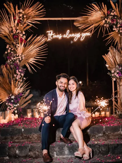 The newly engaged couple celebrating with sparklers, seated amidst a carpet of petals and candles. We create picture-perfect romantic proposal settings.