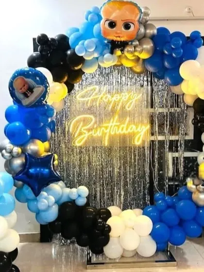 A Boss Baby themed first birthday party with a circular balloon arch in blue, black, and white. The setup is enhanced with a silver fringe backdrop, a "Happy Birthday" neon sign, and a number '1' foil balloon.