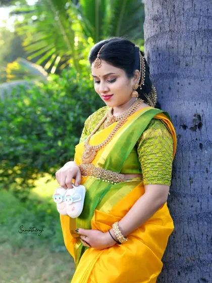 A beautiful outdoor portrait in a traditional yellow saree. The mother-to-be holds a pair of tiny shoes, standing by a palm tree in the warm, natural light.
