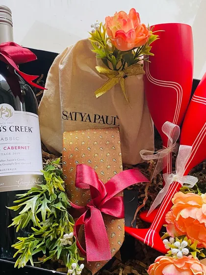A sophisticated gift set perfect for a groomsman or as a couple's gift. It includes a bottle of Shiraz Cabernet, two stylish red wine glasses, and a Satya Paul tie, all elegantly arranged with floral accents in a black gift box.