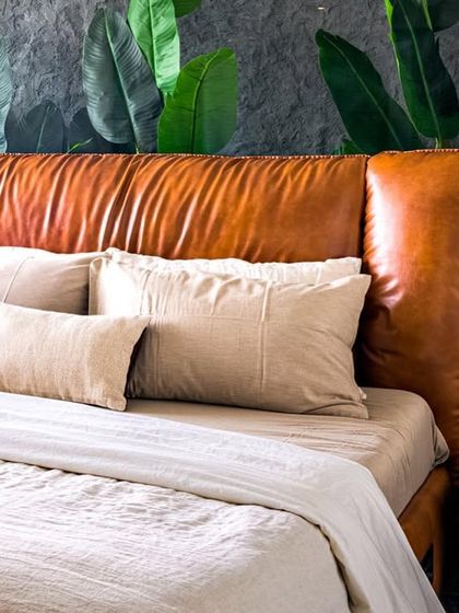 A close-up of the master bedroom in Apartment 56. A plush tan leather headboard is set against a custom mural of tropical leaves, creating a bold yet natural focal point that connects to the green marble of the adjoining bathroom.