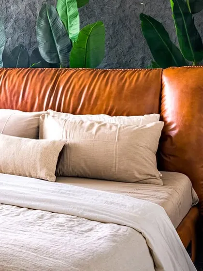 A close-up of the master bedroom in Apartment 56. A plush tan leather headboard is set against a custom mural of tropical leaves, creating a bold yet natural focal point that connects to the green marble of the adjoining bathroom.