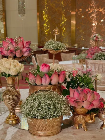 A close-up of a tabletop arrangement, featuring pink lotuses, white roses, and baby's breath in ornate golden vessels. These small details contribute to the overall grandeur.