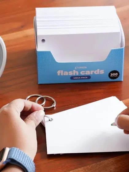 A first-person perspective of someone writing on a flash card at a wooden desk. The cozy setup with a cup of tea and a clock creates a productive and focused learning atmosphere.