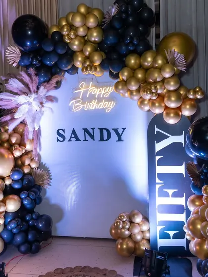 A chic and glamorous 50th birthday celebration setup. The black and gold balloon arch, custom 'Happy Birthday Sandy' backdrop, and 'FIFTY' signage create a sophisticated photo opportunity for a milestone event.