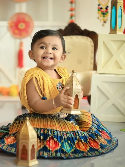 Another joyful shot from a Diwali celebration, this photo shows a baby girl with a huge smile, holding a golden lantern. The festive setup with white blocks and colorful decor adds to the cheerful atmosphere.