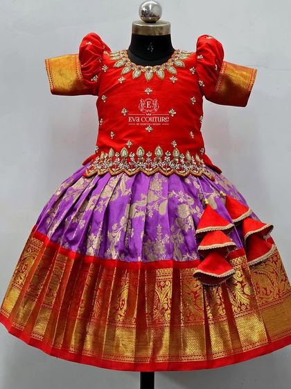 A festive red and purple Kanchi Pattu lehenga. The blouse features a scalloped hem with stone work, and the skirt has a broad golden border for a grand look.
