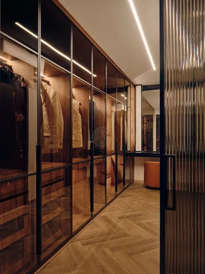 This walk-in closet connects the bedroom and bathroom, featuring dark wood and glass wardrobes with integrated lighting. A built-in vanity table at the end provides a dedicated space for getting ready.