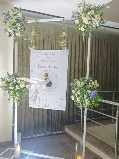We created this beautiful entrance for a baptism ceremony. The setup includes a welcome sign suspended within a white frame, adorned with fresh floral arrangements and candles.
