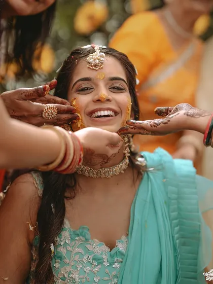 A beautiful candid shot of the bride's joyful expression as her friends and family apply turmeric paste during her Haldi ceremony.