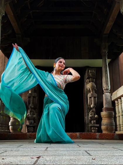 The flowing turquoise saree creates a beautiful sense of movement in this static image, set against the backdrop of traditional Kerala architecture.