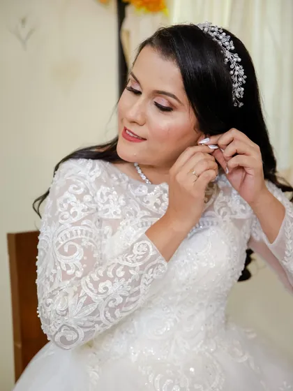 A candid moment of the bride putting on her earrings. Her hair is styled in long, flowing curls, and her makeup is a beautiful soft glam with a hint of shimmer.