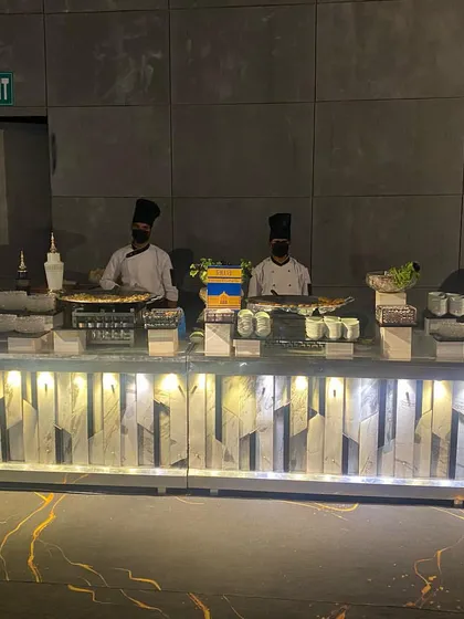 A wide view of our live station spread at an evening event. This setup includes multiple counters where our chefs prepare a variety of dishes, from grilled items to pan-Asian specialties, against a modern, industrial-style backdrop.