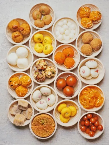 A stunning flat lay of various Indian sweets, each in its own small bowl. This creates a beautiful, grid-like pattern of colors and shapes, showcasing the incredible variety of mithai available.