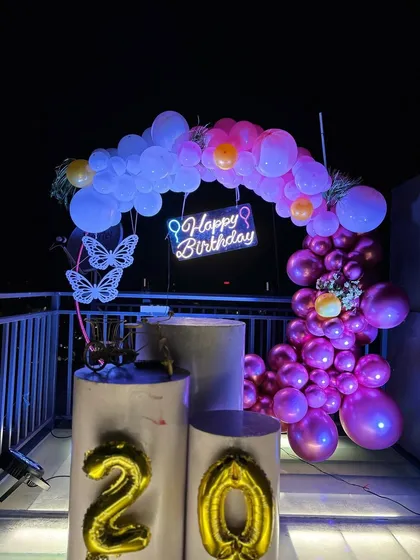 A 20th birthday celebration on a rooftop terrace. The nighttime setup features a balloon arch with a neon "Happy Birthday" sign, illuminated butterfly accents, and foil number balloons.