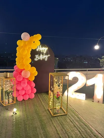 This is a duplicate of ID 21, 60, 67, 108, 121, 168, 206, 232, 239. A vibrant 21st birthday decoration on a rooftop. This setup features a balloon arrangement in hot pink and yellow, a 'Happy Birthday' neon sign, and large marquee numbers against a city skyline.