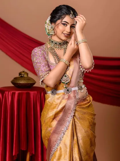 A royal and elegant portrait of a model in a golden-yellow saree. The simple background and classic props like the red-covered table keep the focus entirely on the magnificent outfit and jewelry.