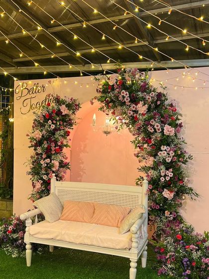 'Better Together'. A romantic garden setup for an intimate celebration, featuring a pink backdrop with a cascading floral arch and a classic white bench, all under a canopy of fairy lights.