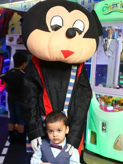 A young child gets a photo with a Mickey Mouse mascot right next to a classic claw machine. The arcade is full of bright lights and exciting characters.