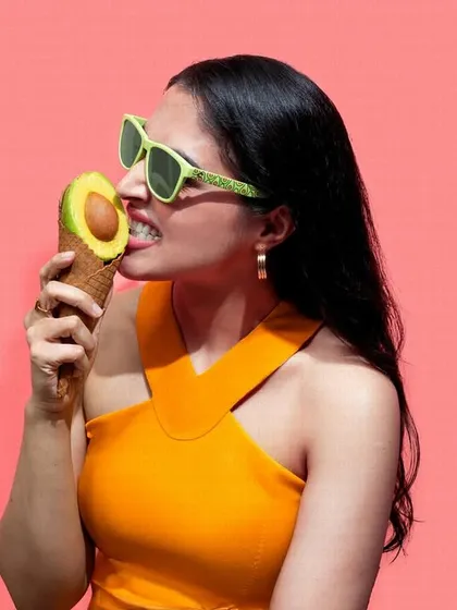 A playful and conceptual shot combining food and fashion. The model "eating" an avocado ice cream cone while wearing matching green sunglasses creates a quirky and color-coordinated image.