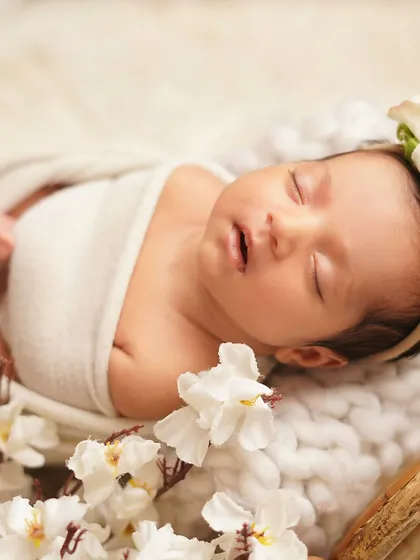 A newborn girl sleeping peacefully, surrounded by white flowers. The soft, angelic styling with a floral headband creates a truly timeless and beautiful infant portrait.