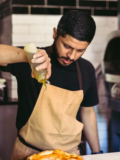 Watch Chef Sudeep in action as he crafts culinary masterpieces, adding a final drizzle of olive oil to a pizza.