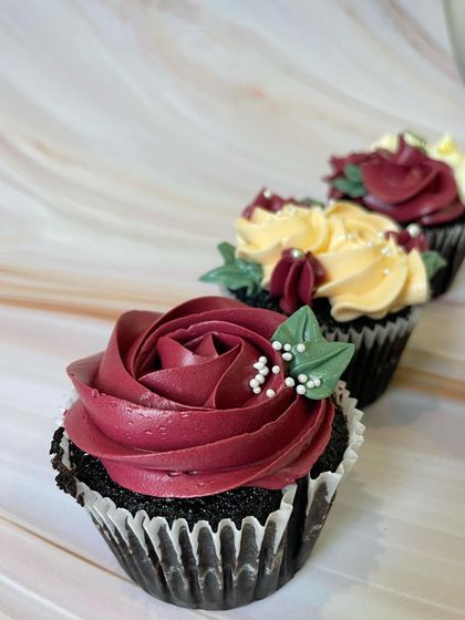 A close-up of a chocolate cupcake with a beautifully piped burgundy buttercream rose. The small green leaves and white sprinkles add a realistic and delicate touch to this mini dessert.