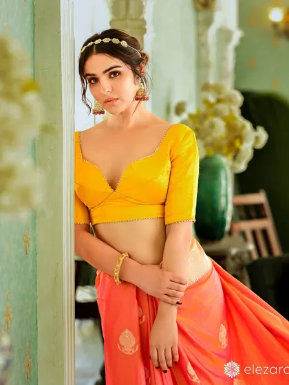 A model for Elezara looking radiant in a bright yellow blouse and orange saree. The soft lighting and her relaxed pose create a warm and inviting image.