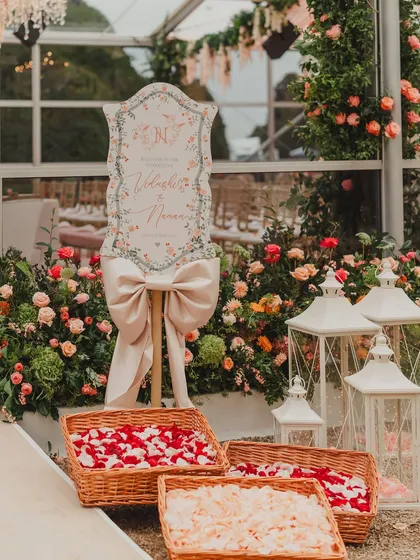 A welcome sign with a large bow, surrounded by lanterns and baskets of flower petals for guests. We believe in creating a warm and beautiful welcome for your guests from the moment they arrive.