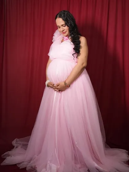 A full length view of the elegant pink maternity gown against our classic red backdrop. This pose beautifully showcases the flowing fabric of the dress and the client's baby bump, creating a timeless memory.