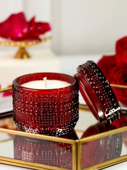 Happy Valentine's Day. This red candle is placed on a mirrored tray, surrounded by deep red roses. The reflection and rich colors create a dramatic and luxurious image perfect for the occasion.