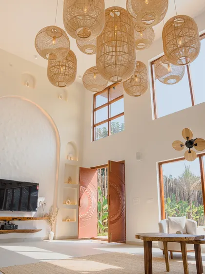 The living area is designed for openness, with double-height ceilings and massive windows framing the tropical landscape. The cluster of woven pendant lights acts as a sculptural centerpiece, enhancing the room's scale and natural aesthetic.